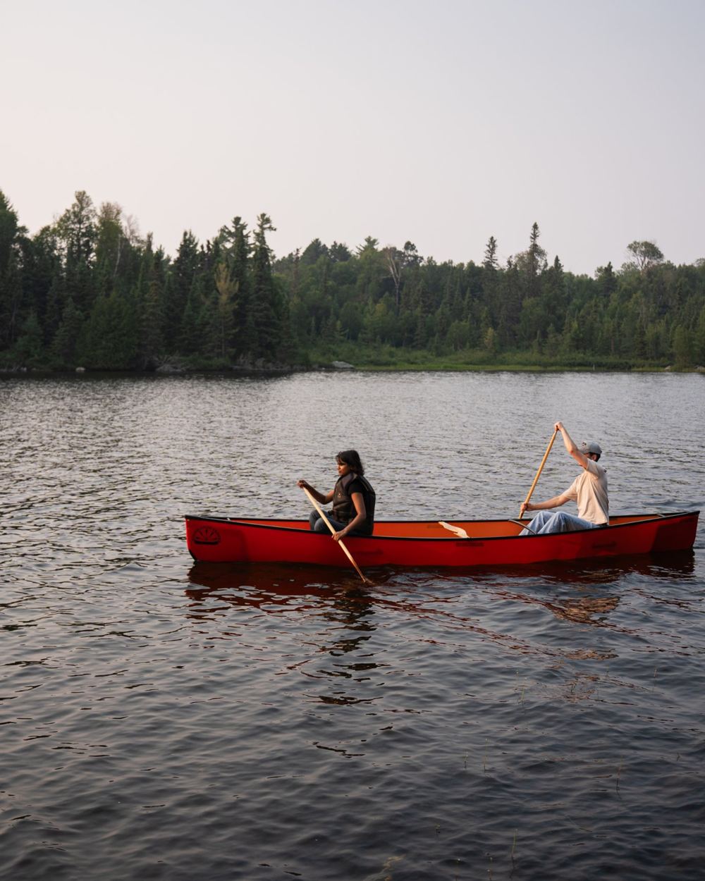 Two people paddling a red canoe past a forested shoreline