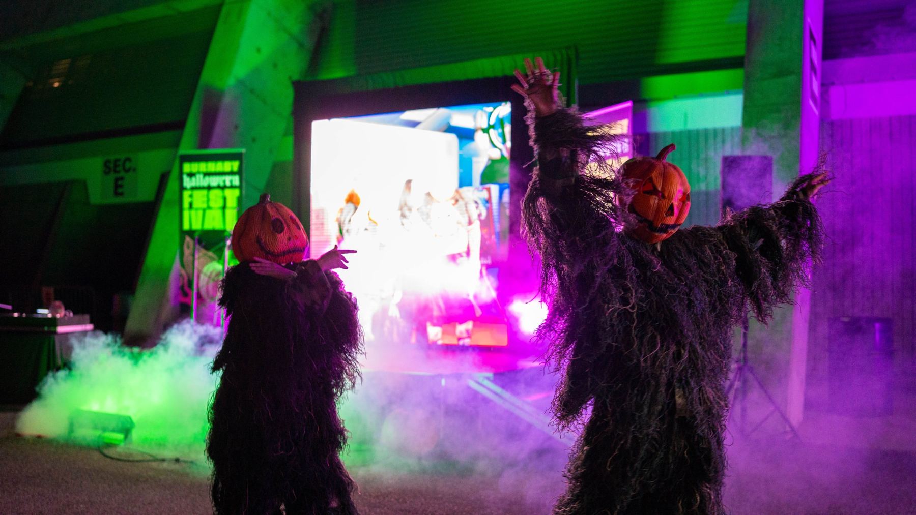 Costumed pumpkin-headed performers dancing in purple and green fog at the festival
